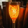A solar-powered LED garden torch with a flame effect, set amongst wooden posts in an outdoor setting. Náhled obrázku produktu