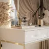 A close-up view of a white vanity table with gold accents, featuring decorative items like a candle, perfume bottle, and makeup brushes. Náhľad obrázku produktu