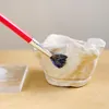 A paintbrush with red handle and dark bristles is applying a clear liquid to a white, textured bowl. A clear rectangular block is visible to the left. Produktbild-Vorschau
