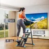 A woman is exercising on a stepper machine in a living room with a large TV screen in the background. Náhled obrázku produktu