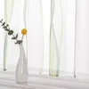 A close-up of a white vase with a single green sprig and a yellow flower, placed on a white shelf in front of sheer curtains with green wavy stripes. Produktbild-Vorschau