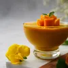 A close-up of a glass bowl filled with mango puree, topped with diced mango and mint leaves. A yellow flower is placed to the left of the bowl. Produktbild-Vorschau