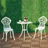 A white metal bistro set with two chairs and a table is placed on a wooden deck in front of a green artificial grass wall. Three bottles of beer are on the table. Produktbild-Vorschau