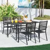 A black metal dining set with four chairs and a table is set up on an outdoor patio, with lush greenery and a distant cityscape in the background. Produktbild-Vorschau