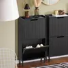A black shoe cabinet with two flip-down drawers is shown in a room with a white chair and a lamp. Náhľad obrázku produktu