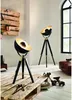Two tripod floor lamps with black shades and brass accents are placed in a room with a wooden pallet wall feature and a large arched window. Podgląd zdjęcia produktu