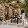 An outdoor patio dining area with a table and chairs set up under a pergola covered in greenery and string lights. Produktbild-Vorschau