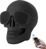 A black skull-shaped Bluetooth speaker with sunglasses, shown with a hand holding a smartphone connected wirelessly. Produktbild-Vorschau