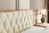 A close-up view of a tufted beige velvet headboard with a wooden trim, next to a bedside table with a lamp. Produktbild-Vorschau