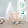 A baby crib with a canopy and bedding, set up in a room with a light blue wall and curtains. Produktbild-Vorschau