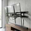 A TV wall mount holding a large flat-screen TV, mounted on a concrete wall above a wooden media console. Náhľad obrázku produktu