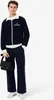 A man wearing a navy blue tracksuit with white trim and a white collar, holding a black bag. Anteprima dell’immagine prodotto