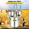 A stainless steel alcohol still is displayed prominently in the foreground, with a backdrop of a golden wheat field and a blue sky. Several bottles of alcoholic beverages and glasses of drinks are arranged on a wooden surface in the lower part of the image Náhled obrázku produktu