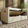 A beige upholstered sofa bed with its storage compartment open, revealing the wooden slatted base and stored items. A charging phone is visible in a side pocket. Produktbild-Vorschau