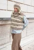 A woman wearing a knitted vest, scarf, and hat, standing in front of a textured wall. Produktbild-Vorschau