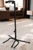 A black guitar stand is shown in a living room setting with a sofa and a side table in the background. Náhľad obrázku produktu