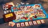 A board game and its components are laid out on a textured surface, possibly outdoors. Produktbild-Vorschau