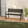 A black metal garden bench is placed on a tiled patio next to a wall, with grass in the foreground and a potted plant to the right. Produktbild-Vorschau