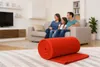 A family is sitting on a couch watching TV in a living room. A rolled-up red blanket is in the foreground. Produktbild-Vorschau