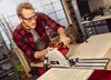 A man is using a circular saw with a guide rail to cut a piece of wood in a workshop. Produktbild-Vorschau