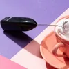 A black milk frother is positioned over a pink cup filled with whipped cream, set against a purple and pink background. Produktbild-Vorschau