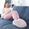 A woman is sitting on a blue couch reading a book while her feet are in a fluffy gray foot warmer. Produktbild-Vorschau