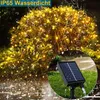 A solar-powered string light set is shown wrapped around a bush, with rain falling and reflecting on the ground. A close-up of the solar panel is inset in the lower right corner. Náhled obrázku produktu