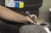 A person's arm and hand are visible, holding a black marker and drawing on a tire. Several tires are stacked in the background, with a Michelin label visible on one. Anteprima dell’immagine prodotto