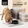 A dog is eating from a pink bowl placed on a tray next to a black bowl. Behind the dog are transparent storage containers filled with pet food and treats. Produktbild-Vorschau