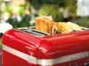 A close-up of a red KitchenAid Artisan toaster with toast and ham inside, set against a blurred green outdoor background. Produktbild-Vorschau