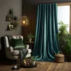 A cozy living room corner with a plush armchair, draped with a dark green throw and cushion, next to a large window covered by dark teal velvet curtains. Several potted plants add a touch of nature to the scene, and a woven basket sits on the floor. Produktbild-Vorschau
