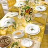 A beautifully set dining table with yellow tablecloth, white plates, cutlery, glasses, a candle, and a centerpiece of vegetables and herbs. Produktbild-Vorschau