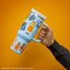 A hand holding a light blue mug with various stickers on it, against an orange background with water splashes. Náhled obrázku produktu