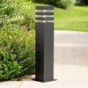 A black garden lamp with clear glass panels stands on a paved walkway next to green bushes. Náhľad obrázku produktu