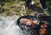 A piece of meat is being fried in a pan over a fire, with vegetables also cooking on the grill. Produktbild-Vorschau