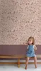 A young child sits on a wooden bench in front of a floral wallpaper. Náhled obrázku produktu