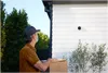 A delivery person wearing a mask and cap holds a package while looking up at a house with a Nest Cam visible on the exterior wall. Voir les images du produit