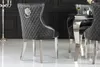 A close-up view of a gray velvet dining chair with a lion head knocker on the back, set against a dark background and a white floor. Produktbild-Vorschau