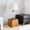A lamp sits on a wooden cube next to a brown ottoman in a room with a concrete wall. Produktbild-Vorschau
