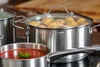A close-up shot of several stainless steel pots on a stovetop, one filled with potatoes and another with tomato soup. Produktbild-Vorschau