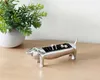 A silver dachshund-shaped jewelry dish sits on a wooden surface next to a potted plant. Podgląd zdjęcia produktu