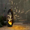A solar-powered garden light with a tree silhouette is placed on a stone patio, casting patterned light on a textured wall. Produktbild-Vorschau