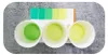 Three small white bowls containing liquids of varying shades of green and yellow, placed in front of a color chart with green and yellow squares. Produktbild-Vorschau