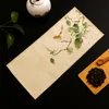 A tea set is laid out on a black surface, with a teapot, a tea mat with a bird and flower design, and a wooden tray with tea leaves and a scoop. Produktbild-Vorschau