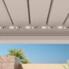 A close-up view of the underside of a white pergola roof, showing the slats and mounting hardware. In the background, a blurred view of a sunny outdoor scene with palm trees and stone structures. Náhled obrázku produktu