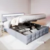 A grey upholstered bed with its storage compartment open, revealing pillows and a decorative flower. Voir les images du produit