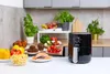 A black air fryer sits on a kitchen counter next to various food items. Produktbild-Vorschau