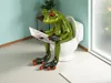 A decorative frog figurine sitting on a toilet and reading a newspaper in a bathroom. Produktbild-Vorschau