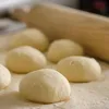 Several balls of dough are arranged on a surface dusted with flour, with a wooden rolling pin in the background. Produktbild-Vorschau