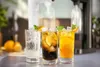 Three glasses of sparkling drinks with ice, lemon and mint garnishes, on a counter with a soda maker in the background. Produktbild-Vorschau